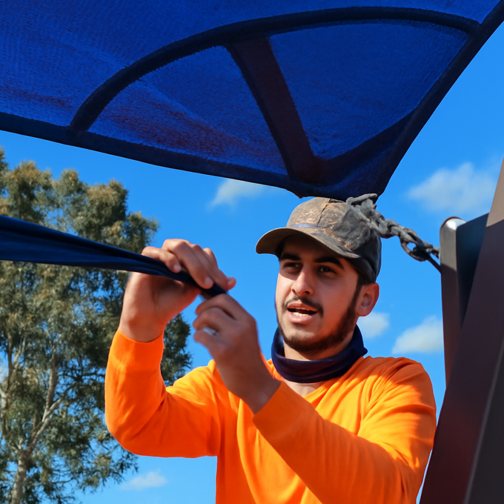 Worker tightening blue shade sail using rope and carabiner in a park setting.
