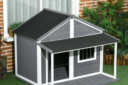 Large outdoor dog house with porch roof placed in a garden near a brick home.
